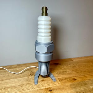 Oversized spark plug lamp with the white ceramic section designed as a light shade. Bronze top section, white shade and silver base. With a white cable. Stood on a wooden desk and grey background.