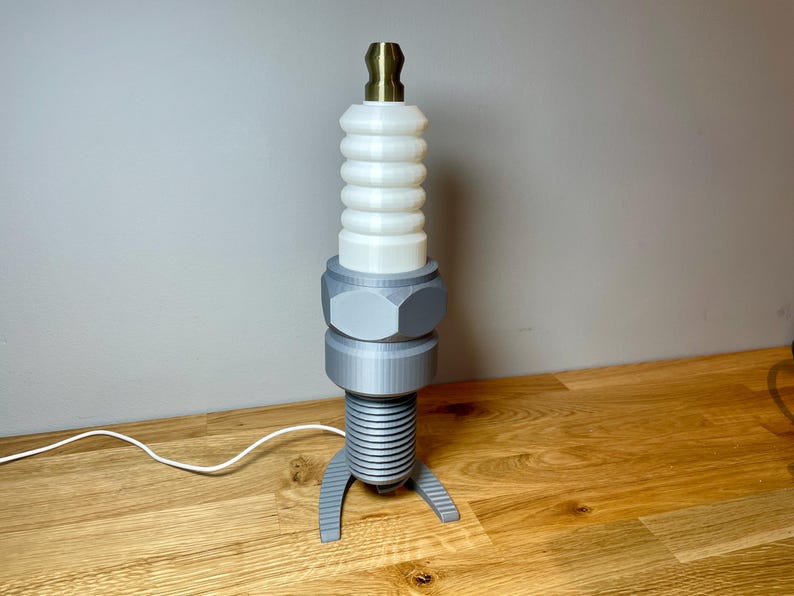 Oversized spark plug lamp with the white ceramic section designed as a light shade. Bronze top section, white shade and silver base. With a white cable. Stood on a wooden desk and grey background.