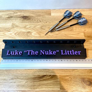 Puede incluir: Un soporte de dardos negro con el texto "Luke 'The Nuke' Littler" en morado. Tres dardos con ca&ntilde;as negras y plateadas y plumas azules POWER est&aacute;n sobre una superficie de madera. Tambi&eacute;n hay una regla.