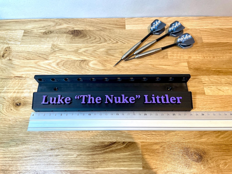 Puede incluir: Un soporte de dardos negro con el texto "Luke 'The Nuke' Littler" en morado. Tres dardos con ca&ntilde;as negras y plateadas y plumas azules POWER est&aacute;n sobre una superficie de madera. Tambi&eacute;n hay una regla.