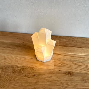 Pu&ograve; includere: Una lampada bianca a forma di cristallo con una luce calda, appoggiata su una superficie di legno.