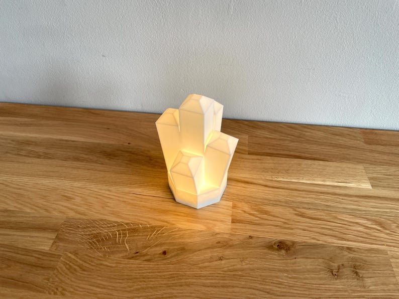 Pu&ograve; includere: Una lampada bianca a forma di cristallo con una luce calda, appoggiata su una superficie di legno.