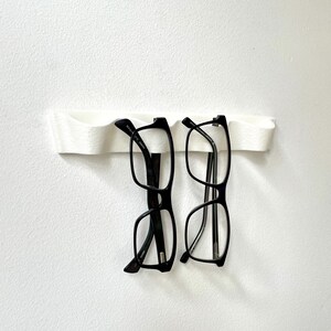 May include: A white, wall-mounted eyeglasses holder with two pairs of black-framed glasses. The holder has a curved design, and the glasses are suspended from it. The background is a plain white wall.