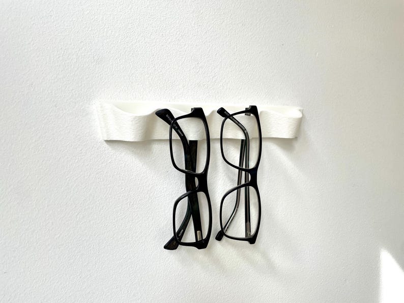 May include: A white, wall-mounted eyeglasses holder with two pairs of black-framed glasses. The holder has a curved design, and the glasses are suspended from it. The background is a plain white wall.