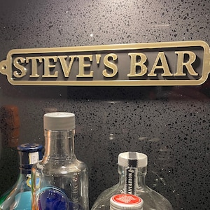 May include: A gold-colored metal sign with the text "STEVE'S BAR" in black lettering. The sign is mounted on a black surface.