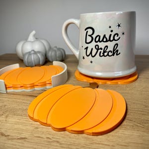 Puede incluir: Una taza de cer&aacute;mica blanca con el texto "Basic Witch" y gr&aacute;ficos de estrellas se encuentra sobre un posavasos naranja con forma de calabaza. Posavasos de calabaza naranja adicionales est&aacute;n en un soporte blanco, con calabazas blancas y grises decorativas en el fondo.