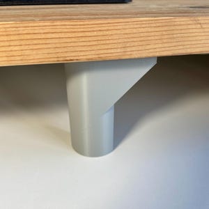 May include: A close-up of a light gray wooden desk with a white plastic leg. The desk is made of wood with a natural finish. The leg is made of plastic and has a simple design.