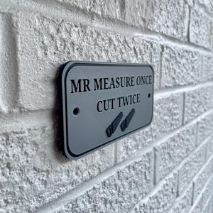 May include: A rectangular gray sign with black trim is mounted on a white brick wall. The sign reads "MR MEASURE ONCE CUT TWICE" with two saw illustrations. The sign has two small holes for mounting.