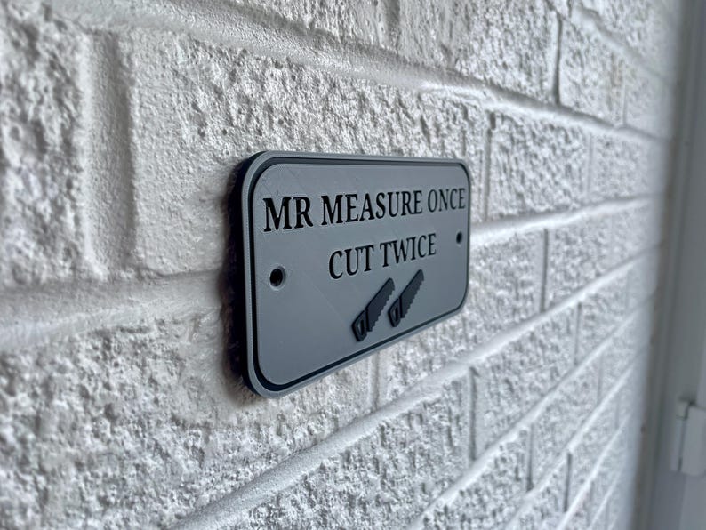 May include: A rectangular gray sign with black trim is mounted on a white brick wall. The sign reads "MR MEASURE ONCE CUT TWICE" with two saw illustrations. The sign has two small holes for mounting.