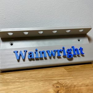 Wall mounted dart holder. Silver with Blue personalised text on the front reading Wainwright