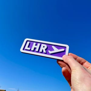 May include: Purple sign with white lettering that reads "LHR" and a white airplane icon pointing to the right.