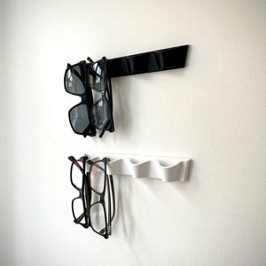 May include: Two wall-mounted eyeglass holders. The top holder is black and holds two pairs of sunglasses. The bottom holder is white and holds two pairs of eyeglasses. The holders are mounted on a white wall.