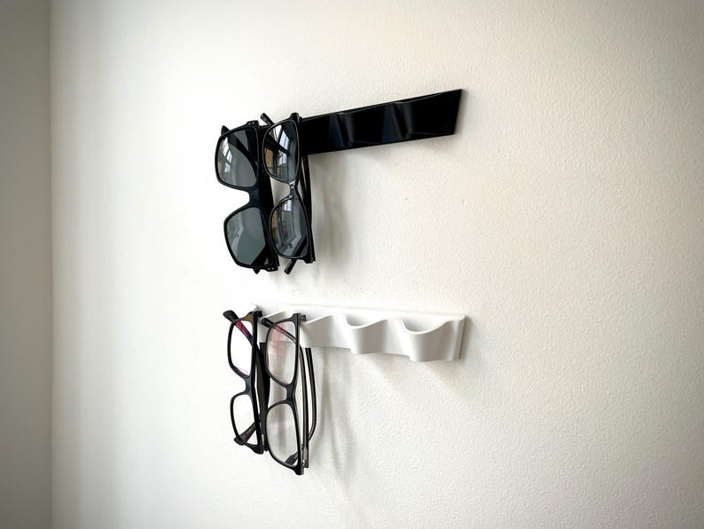May include: Two wall-mounted eyeglass holders. The top holder is black and holds two pairs of sunglasses. The bottom holder is white and holds two pairs of eyeglasses. The holders are mounted on a white wall.