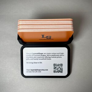 May include: A black case holds a stack of orange and white cards. The top card has the text "LS LAYEREDSHAPE". The case also displays text: "Here at LayeredShape...We bring ideas to life." and a QR code. Visit layeredshape.etsy.com.