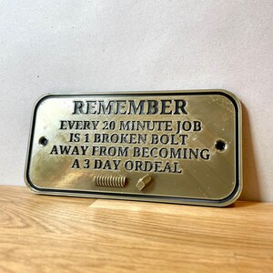 May include: A gold-colored metal sign with black text that reads "REMEMBER EVERY 20 MINUTE JOB IS 1 BROKEN BOLT AWAY FROM BECOMING A 3 DAY ORDEAL".