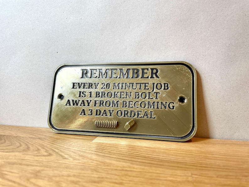 May include: A gold-colored metal sign with black text that reads "REMEMBER EVERY 20 MINUTE JOB IS 1 BROKEN BOLT AWAY FROM BECOMING A 3 DAY ORDEAL".