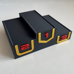 May include: A black three-tiered victory podium with a raised central platform. The front of each tier features a gold-colored U-shaped border surrounding a red number. The numbers 2, 1, and 3 are displayed from left to right on the respective tiers.