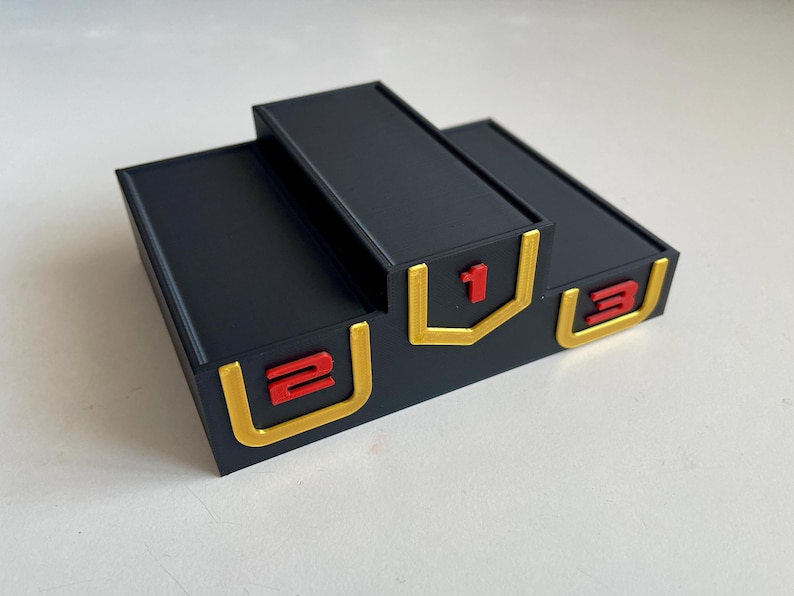 May include: A black three-tiered victory podium with a raised central platform. The front of each tier features a gold-colored U-shaped border surrounding a red number. The numbers 2, 1, and 3 are displayed from left to right on the respective tiers.