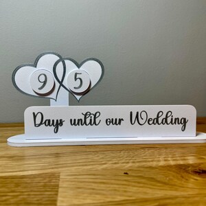 May include: White wooden countdown sign with two intertwined hearts, each with a number on it. The sign reads "Days until our Wedding".