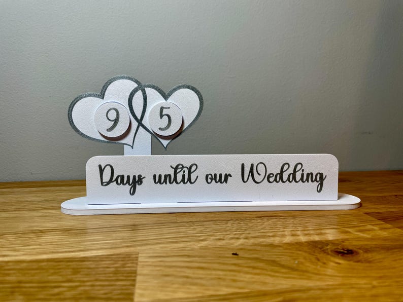 May include: White wooden countdown sign with two intertwined hearts, each with a number on it. The sign reads "Days until our Wedding".