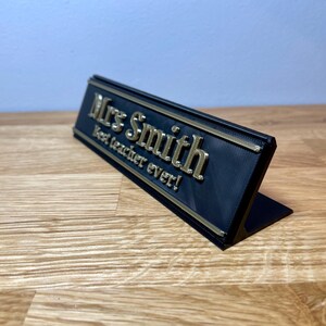 name plaque / plate with a teachers name and best teacher ever! text on the front. Black with bronze text on a wooden desk with grey background