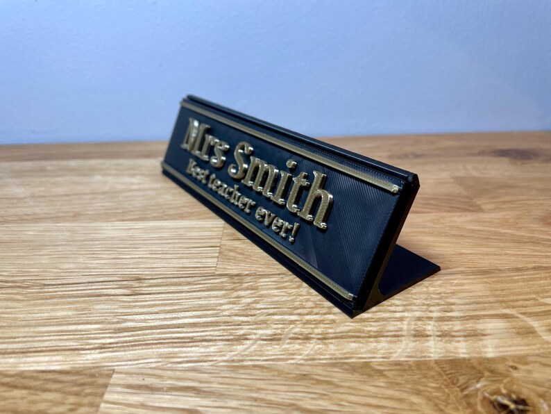 name plaque / plate with a teachers name and best teacher ever! text on the front. Black with bronze text on a wooden desk with grey background