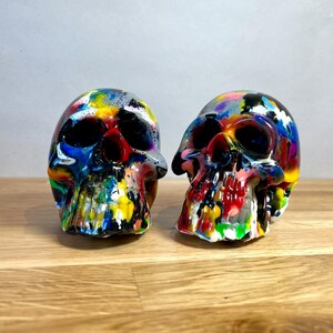 May include: Two colourful, hand-painted skull figurines. The skulls are painted in a variety of bright colours, including red, yellow, blue, green, and purple. The skulls are made of a smooth, white material and have a glossy finish.