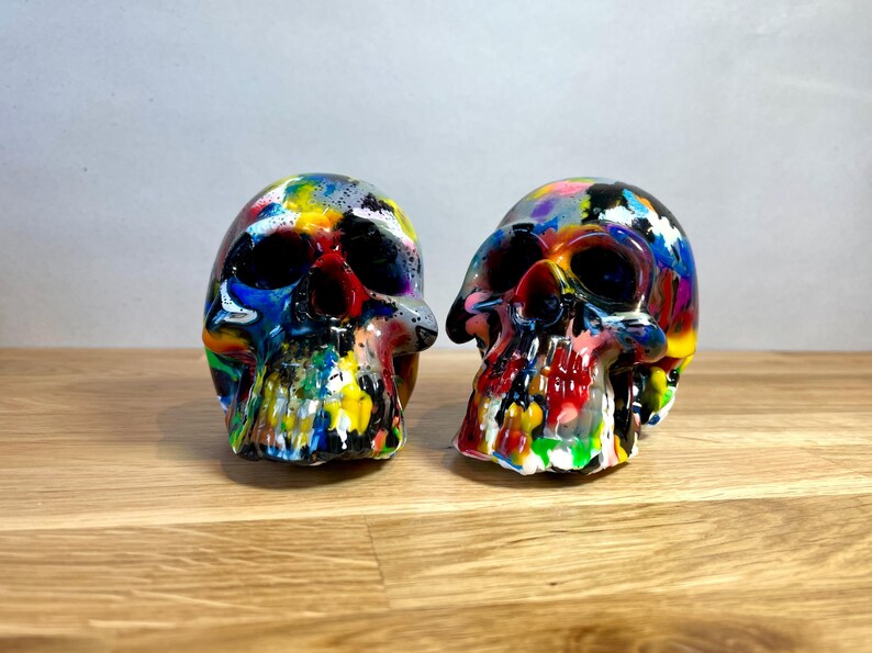 May include: Two colourful, hand-painted skull figurines. The skulls are painted in a variety of bright colours, including red, yellow, blue, green, and purple. The skulls are made of a smooth, white material and have a glossy finish.