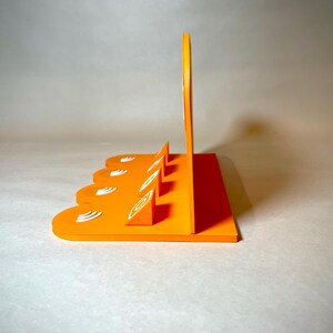 May include: An orange, geometric display stand with a series of angled slots and a tall, curved back. The stand features white, stylized leaf-like designs. The design is modern and minimalist, suitable for displaying items.