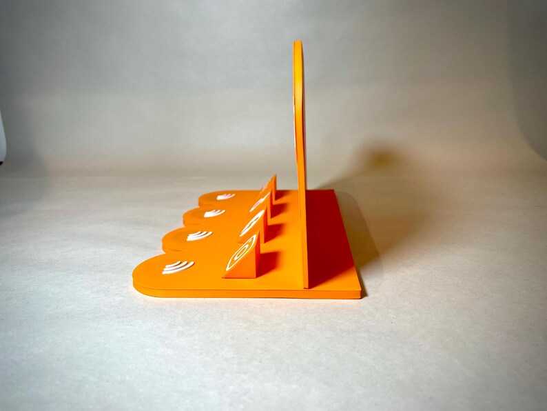May include: An orange, geometric display stand with a series of angled slots and a tall, curved back. The stand features white, stylized leaf-like designs. The design is modern and minimalist, suitable for displaying items.