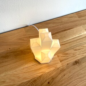 Pu&ograve; includere: Una lampada geometrica bianca con un design moderno. La lampada &egrave; accesa ed emette una luce calda.