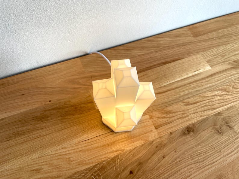 Pu&ograve; includere: Una lampada geometrica bianca con un design moderno. La lampada &egrave; accesa ed emette una luce calda.