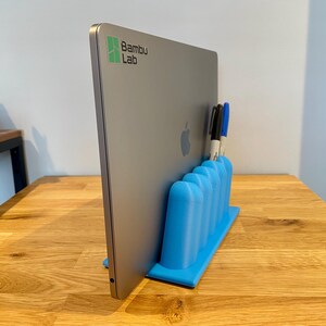 May include: A bright blue 3D-printed vertical laptop stand with five rounded slots for holding pens or markers. A silver laptop with a Bambu Lab logo and an Apple logo is inserted into the stand, which is positioned on a wooden surface.