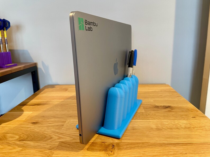 May include: A bright blue 3D-printed vertical laptop stand with five rounded slots for holding pens or markers. A silver laptop with a Bambu Lab logo and an Apple logo is inserted into the stand, which is positioned on a wooden surface.