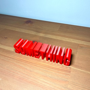 Pu&ograve; includere: La parola "SOMETHING" in rosso vivo, tridimensionale, &egrave; esposta su una superficie di legno. Le lettere a blocchi sono in piedi, proiettando ombre. Lo sfondo &egrave; un muro azzurro.