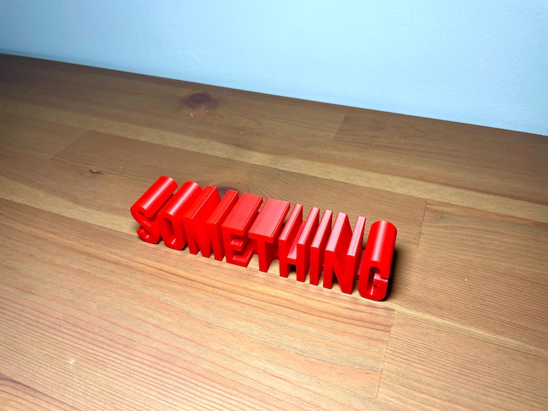 Pu&ograve; includere: La parola "SOMETHING" in rosso vivo, tridimensionale, &egrave; esposta su una superficie di legno. Le lettere a blocchi sono in piedi, proiettando ombre. Lo sfondo &egrave; un muro azzurro.