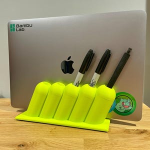 May include: A bright neon yellow desk organizer with five slanted cylindrical slots holding two black markers and one black stylus. The organizer is positioned in front of a silver laptop featuring a Bambu Lab logo and a green Bulbasaur sticker.