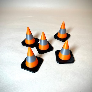 May include: Five miniature orange and grey traffic cones with black square bases are arranged on a light background. The cones have a reflective grey band and are likely used for decoration or model projects.