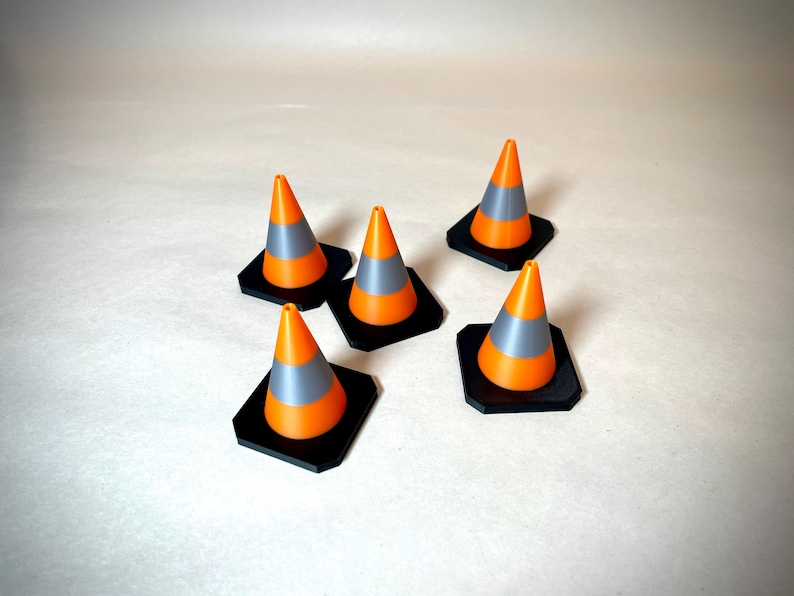 May include: Five miniature orange and grey traffic cones with black square bases are arranged on a light background. The cones have a reflective grey band and are likely used for decoration or model projects.