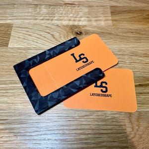 May include: A black and gray patterned card holder with a cutout for a card. The card holder is on top of two orange cards with the text "LAYEREDSHAPE" and the letter "S" in a stylized font.