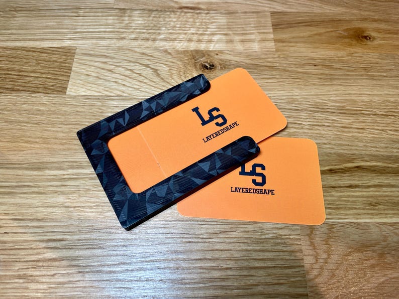 May include: A black and gray patterned card holder with a cutout for a card. The card holder is on top of two orange cards with the text "LAYEREDSHAPE" and the letter "S" in a stylized font.