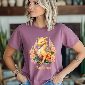May include: A mauve t-shirt with a yellow dragon graphic and floral accents. The dragon is surrounded by pink and yellow flowers. The text "Andarna" is printed in gold below the dragon.