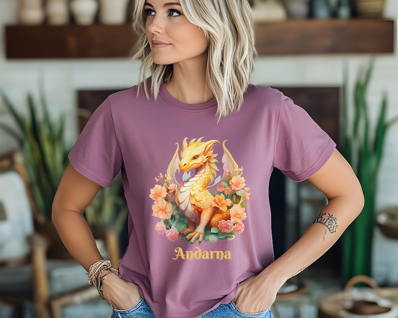 May include: A mauve t-shirt with a yellow dragon graphic and floral accents. The dragon is surrounded by pink and yellow flowers. The text "Andarna" is printed in gold below the dragon.