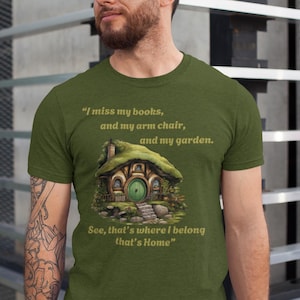 May include: A green t-shirt with a quote about missing books, an armchair, and a garden. The quote is accompanied by an illustration of a small, green, round house with a door and a window. The text on the shirt reads: "I miss my books, and my arm chair, and my garden. See, that's where I belong that's Home."