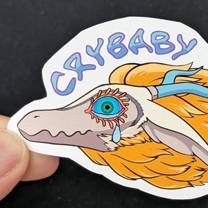 May include: A white and grey cartoon dragon with a blue antler, orange hair, and a single blue eye with a tear. The dragon is crying and the text "CRYBABY" is written above it in blue.