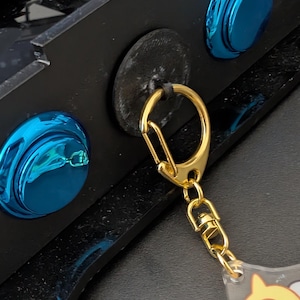 Puede incluir: Un llavero dorado con un dije de plástico transparente que presenta un personaje de dibujos animados. El llavero está unido a una superficie negra con un botón azul.