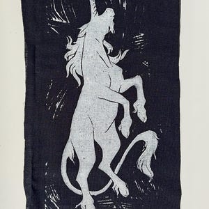 May include: A dark blue fabric featuring a white unicorn design. The unicorn is depicted rearing up on its hind legs. The fabric is rectangular with a raw edge. A safety pin with a small white bead is attached.