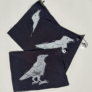 May include: Three black fabric patches featuring white raven designs. One patch depicts a raven's head, another a raven in flight, and the third a standing raven. A safety pin with white beads is attached.