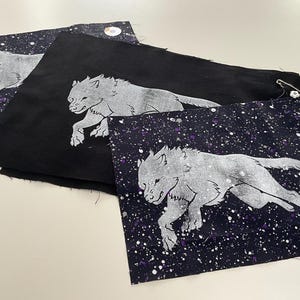 May include: Three rectangular fabric pieces featuring a silver-coloured wolf illustration. Two pieces have a dark purple and black galaxy-like background with white speckles, while the centre piece is solid black. A small safety pin is attached.