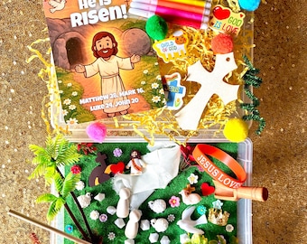 May include: An open clear plastic container filled with Easter-themed craft supplies. Includes a book with the text "He is Risen!", markers, stickers, a wooden cross, and a miniature scene with figures and greenery. The text "Matthew 28, Mark 16, Luke 24, John 20" is also visible.